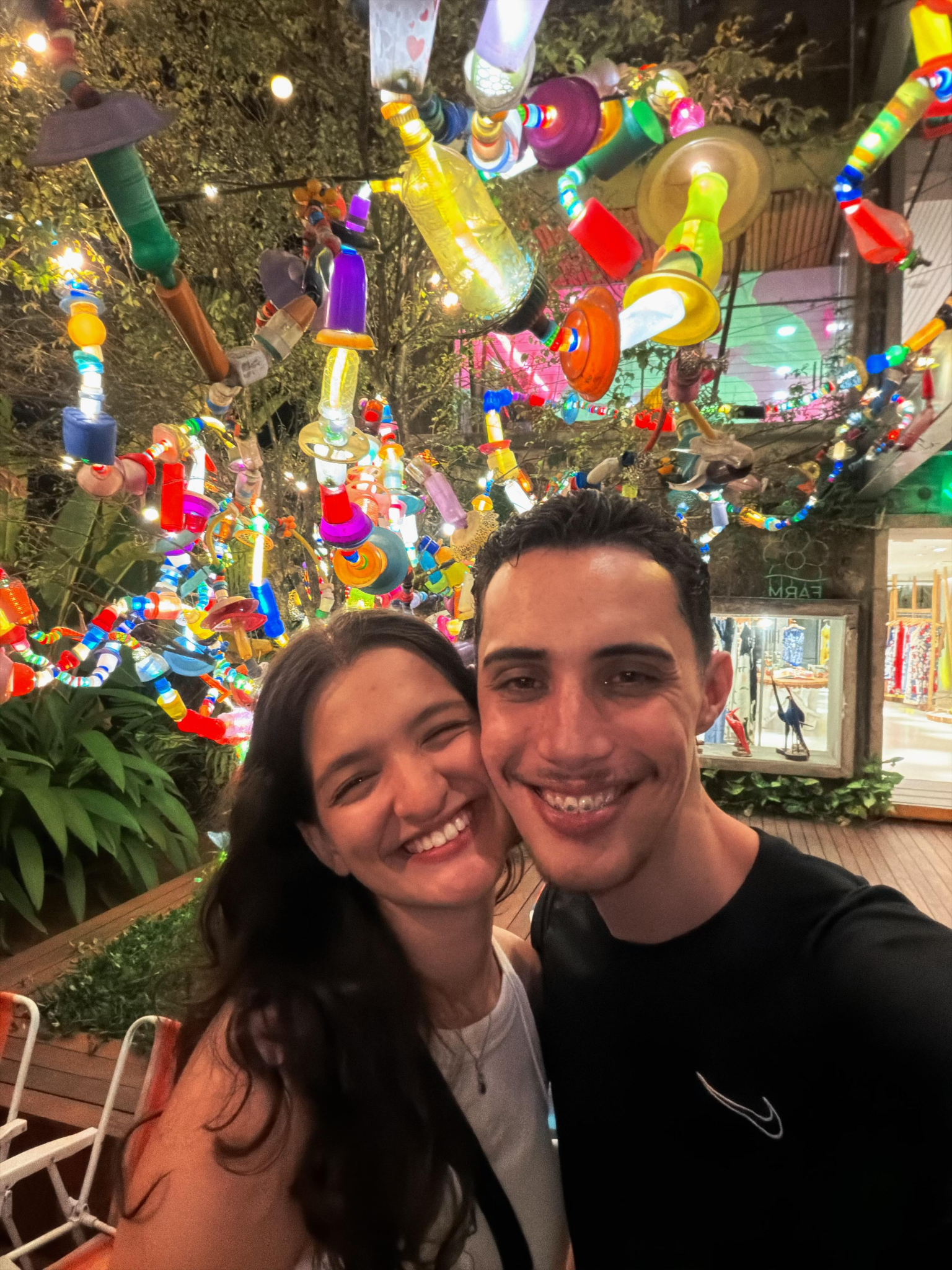 Sophia e Kauê sorrindo sob uma instalação colorida de luzes e objetos de vidro