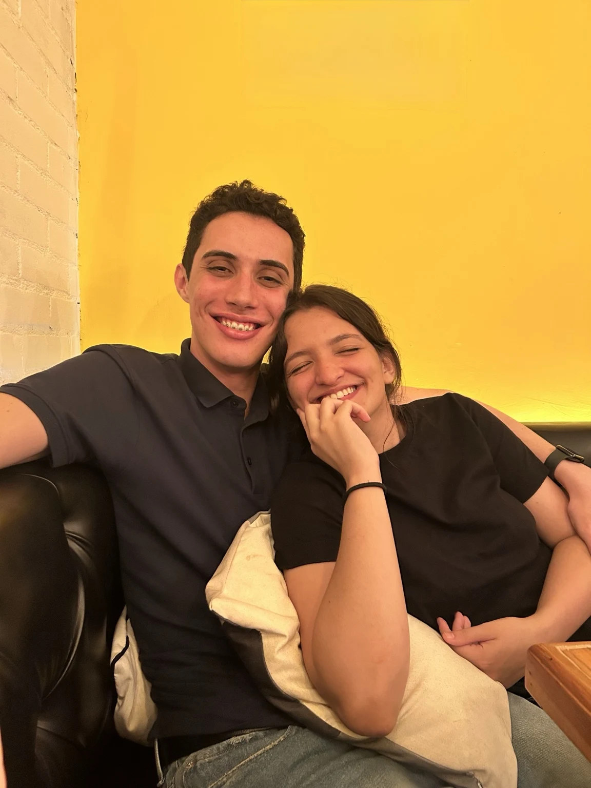 Sophia e Kauê abraçados e sorrindo em frente a uma parede amarela