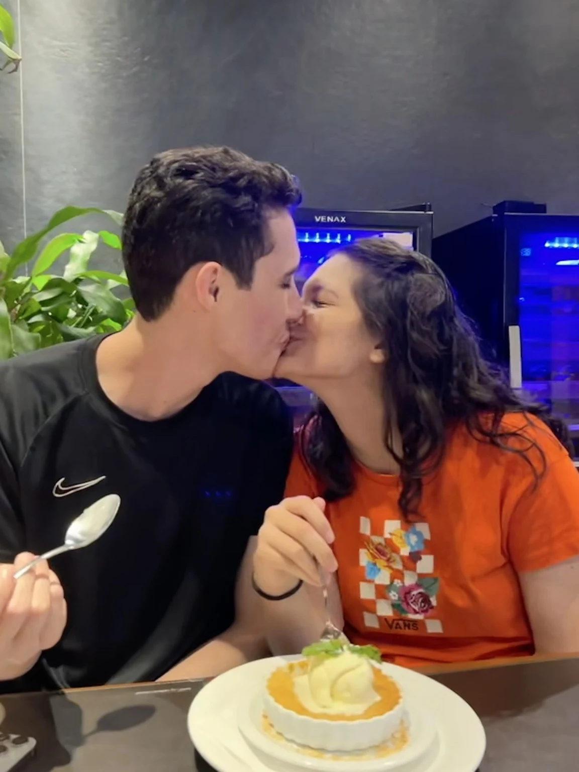 Sophia e Kauê se beijando à mesa, com uma sobremesa